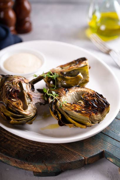 Roasted artichokes garnished with herbs served with a dipping sauce on a white plate.