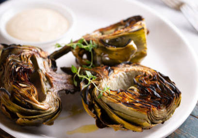 Roasted artichokes garnished with herbs served with a dipping sauce on a white plate.