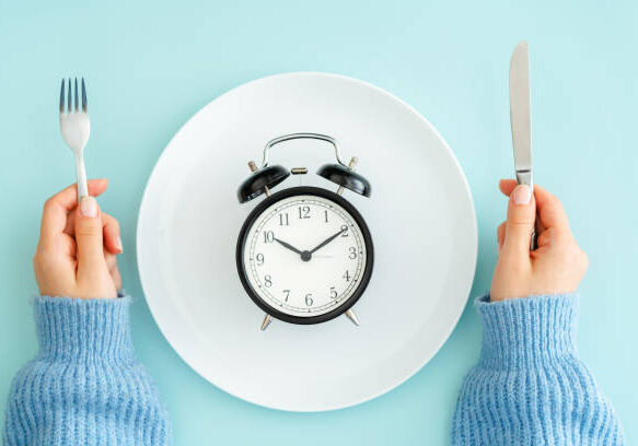 Person holding fork and knife with an alarm clock on a plate, illustrating fasting.