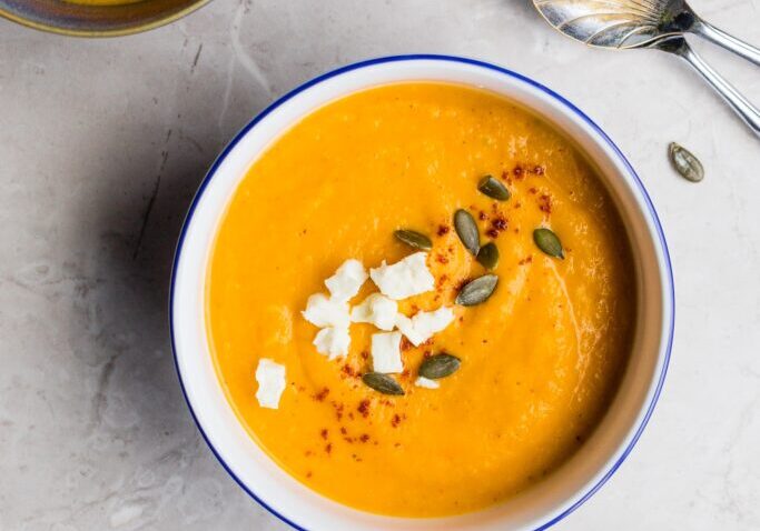 Bowl of vibrant orange vegetable soup garnished with seeds and herbs, promoting gut and hormone heal.