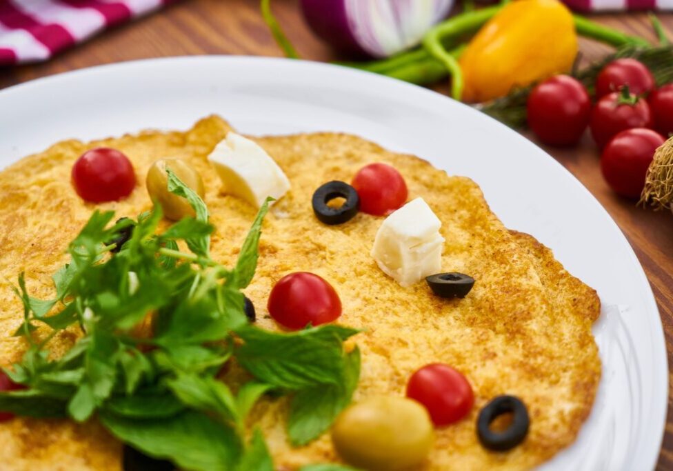 Delicious moong omelette garnished with cherry tomatoes, olives, and herbs on a white plate.
